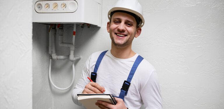 Smiling technician in a hard hat and overalls taking notes during a tankless water heater installation assessment.